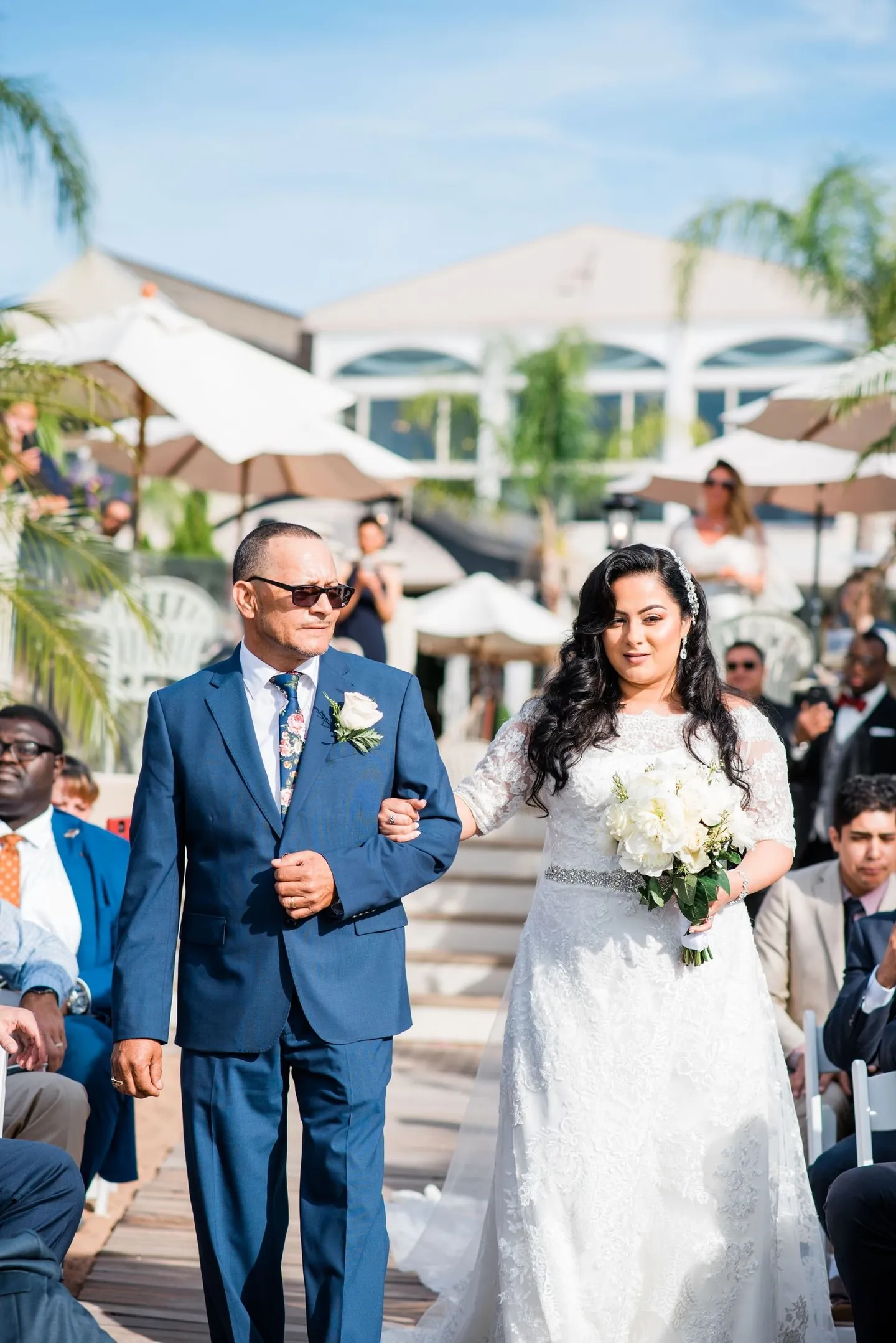 Man in a blue suit walks the bride down the boardwalk aisle past umbrellas