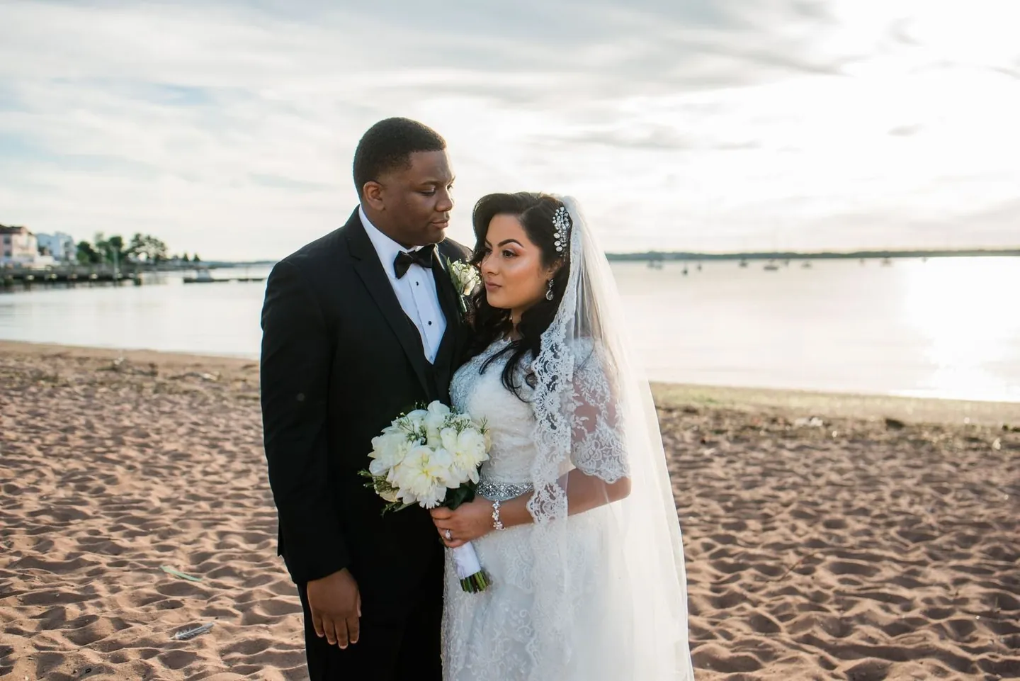Natasha and Jordan stand close on the sand with the calm harbor and far shoreline behind