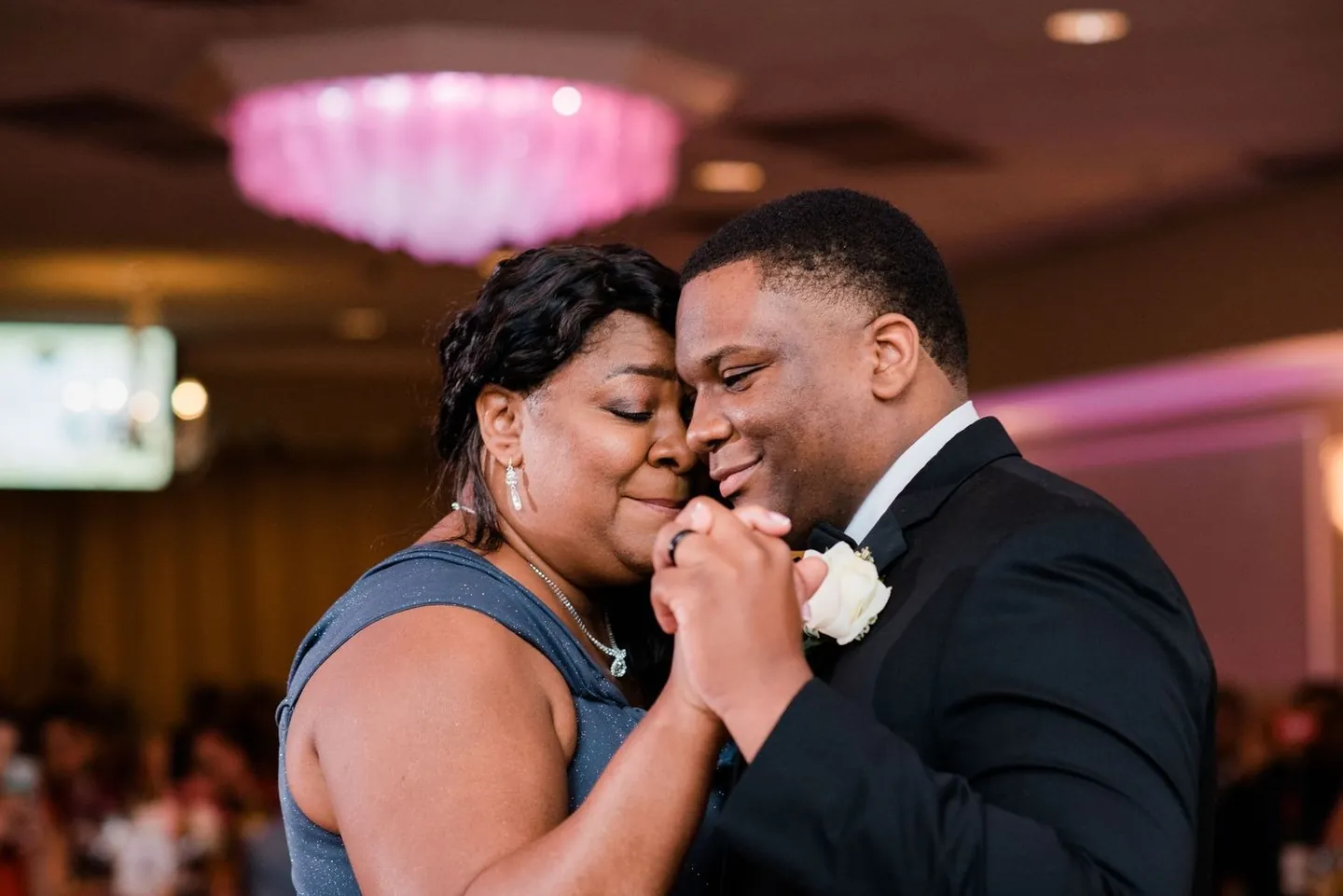 Groom dances cheek to cheek with a woman in a grey dress under pink chandeliers