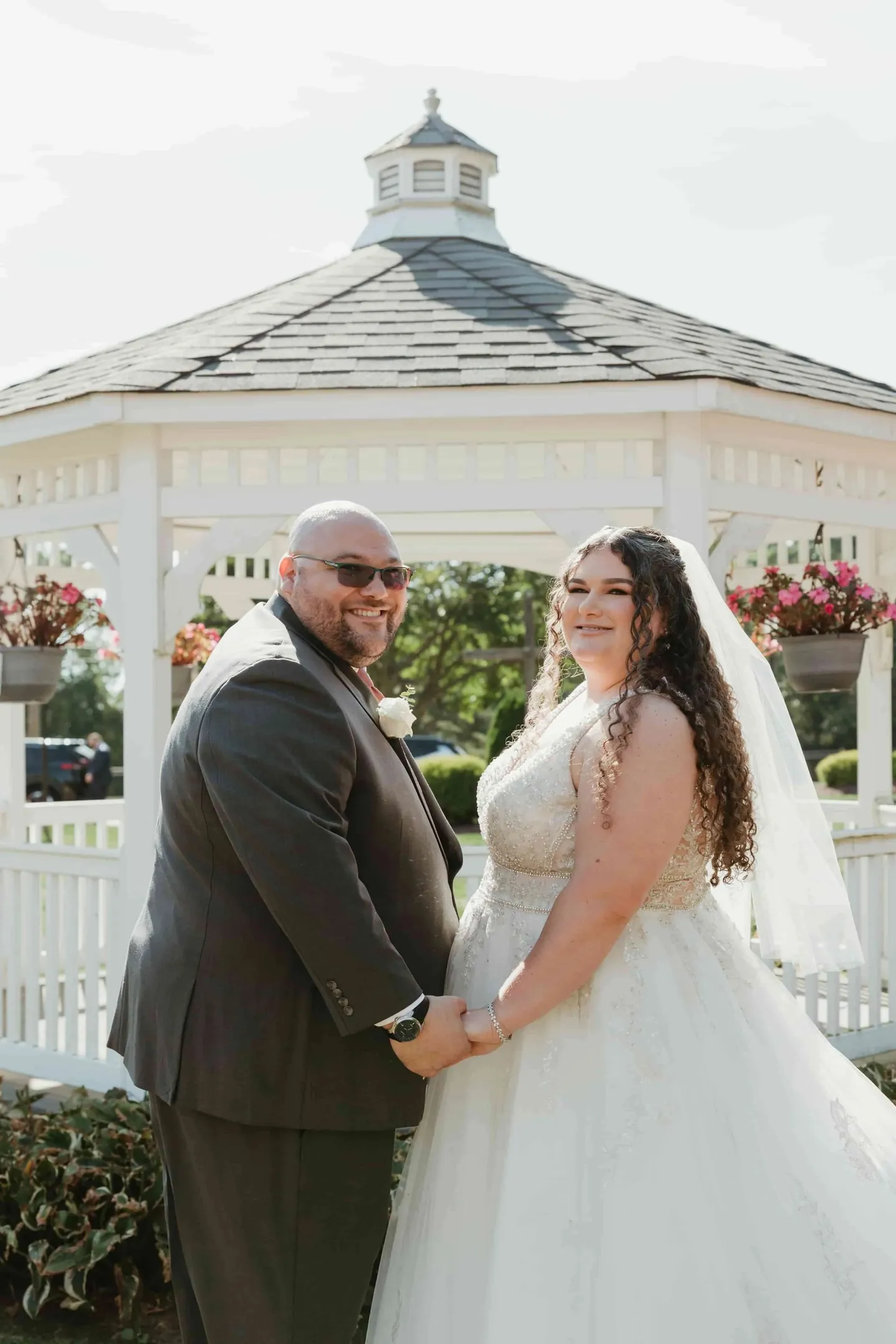 Nicole and Ryan hold hands and smile in front of a white gazebo with hanging flower baskets