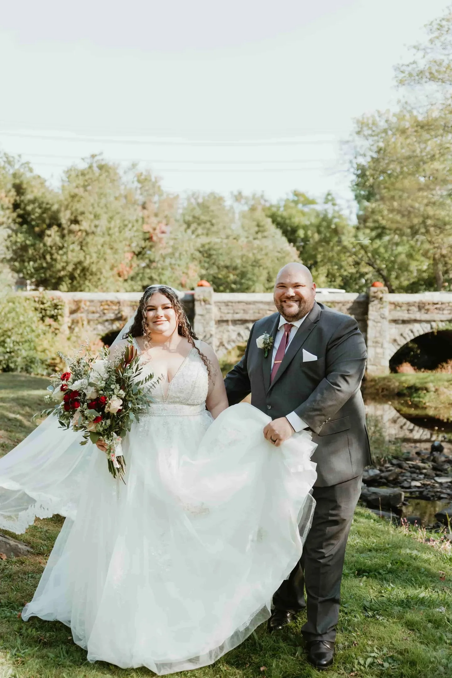 Nicole and Ryan laugh as she lifts her full skirt on the grass near a stone bridge