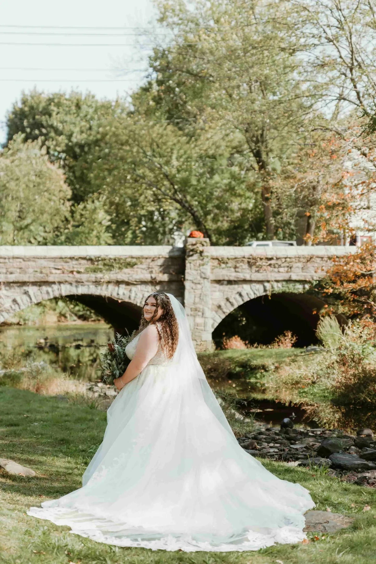 The bride looks back over her shoulder by the stone bridge, her gown spread on the grass
