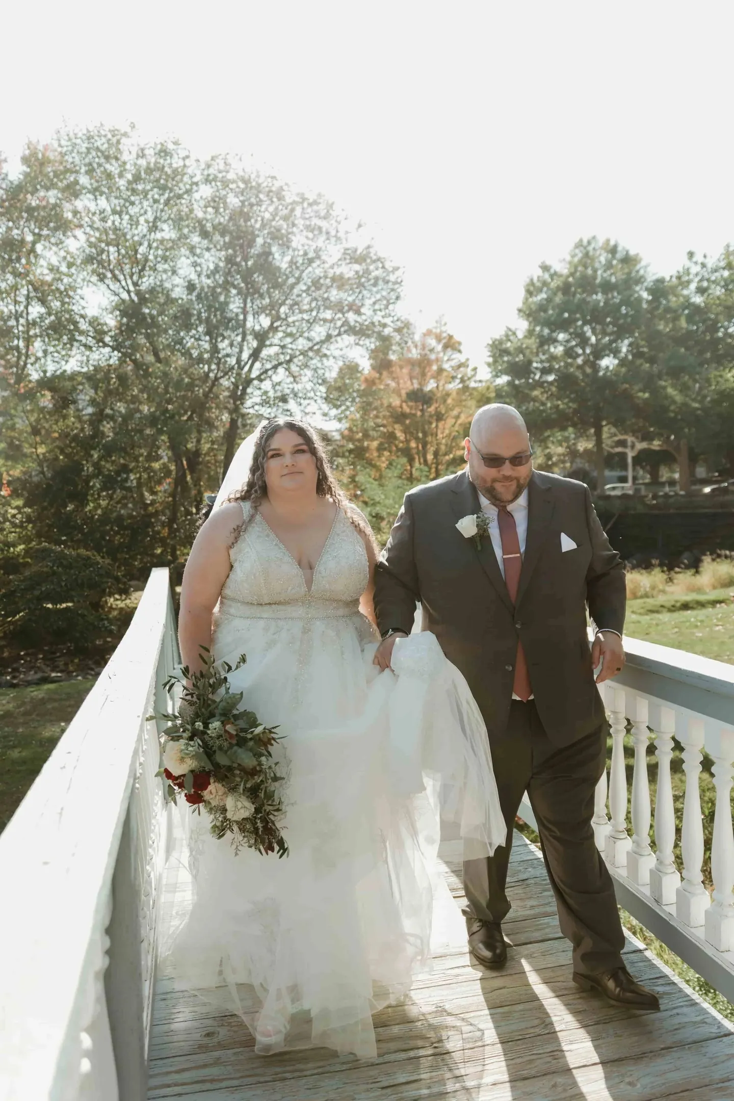 Nicole and Ryan cross a white wooden footbridge together, trees glowing behind them