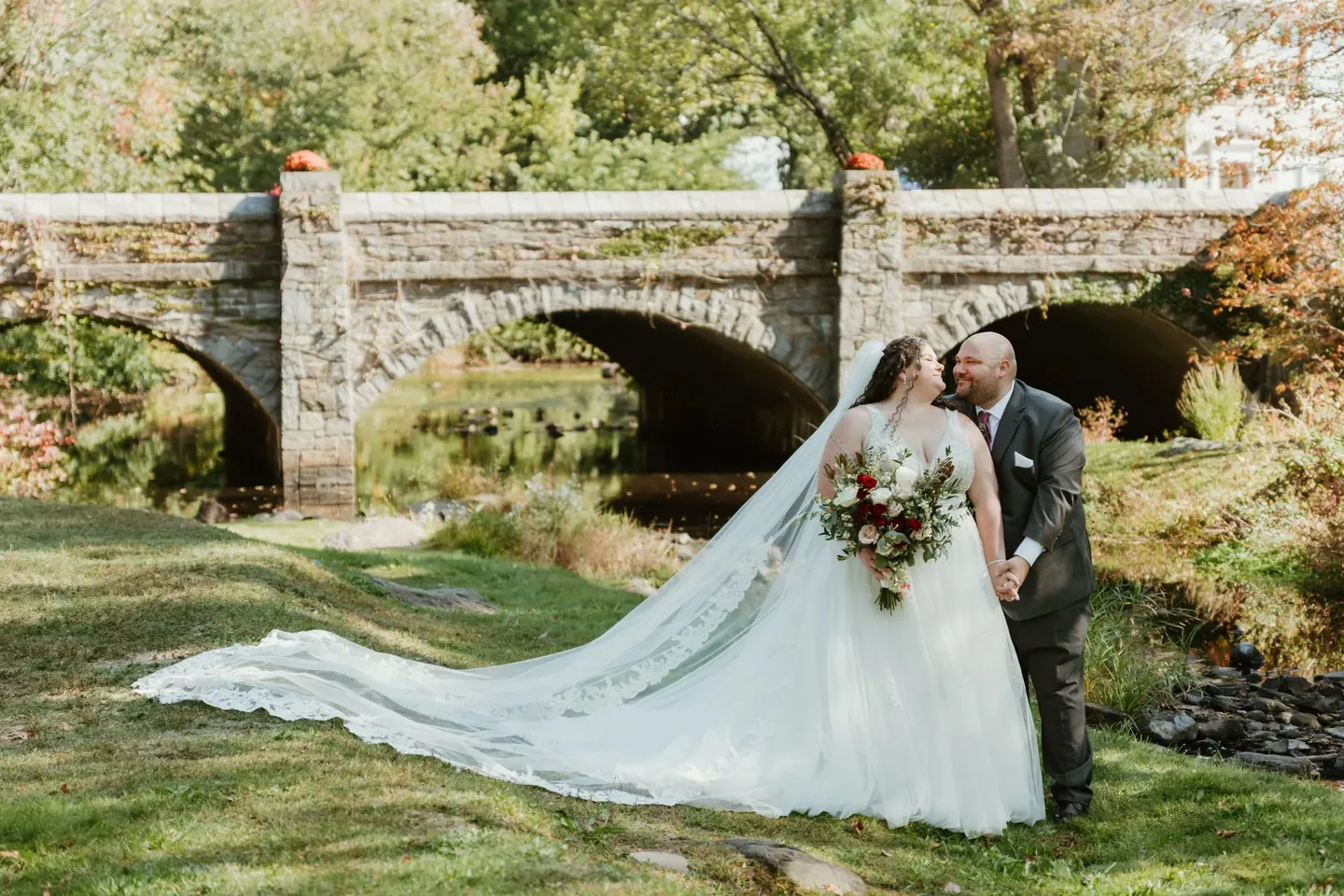 Nicole and Ryan kiss beside an old stone arch bridge, her veil sweeping across the grass