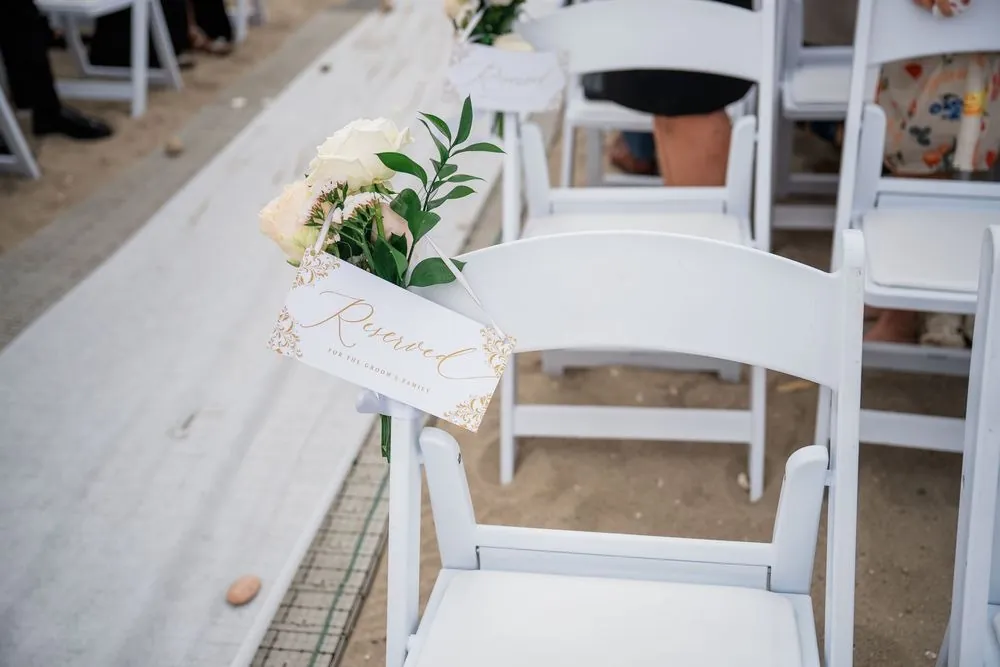 White folding chair on the beach aisle with a gold reserved card and a sprig of greenery