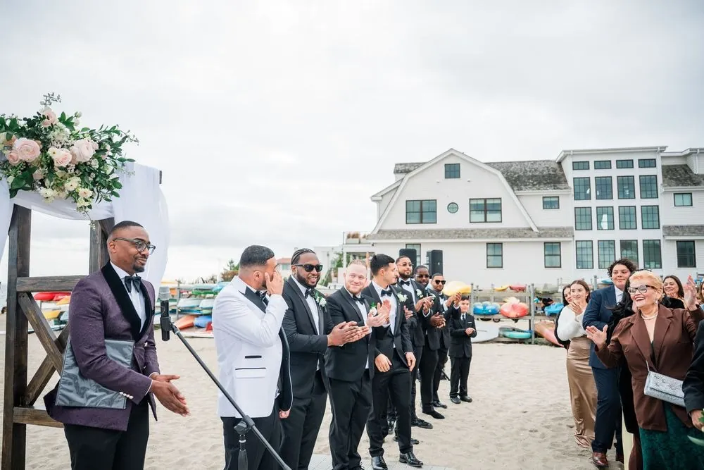 Elvin in a white dinner jacket waits by the floral arch as groomsmen and guests clap on the sand