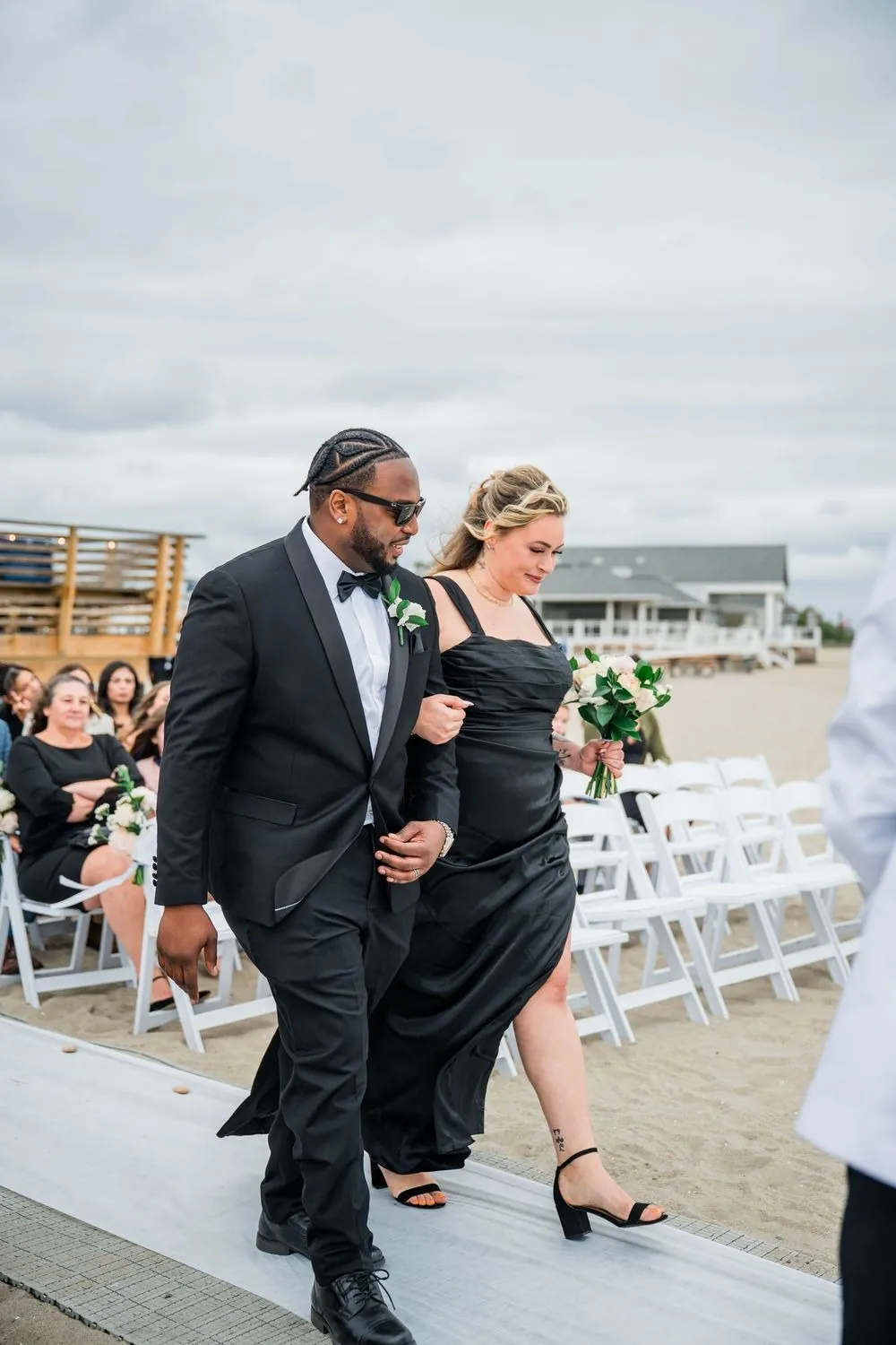 Groomsman in a black tuxedo walks a bridesmaid down the beach aisle past white chairs