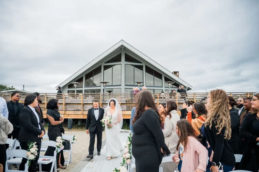 Olivia walks the aisle on an escort's arm with the shingled pavilion and packed deck behind her