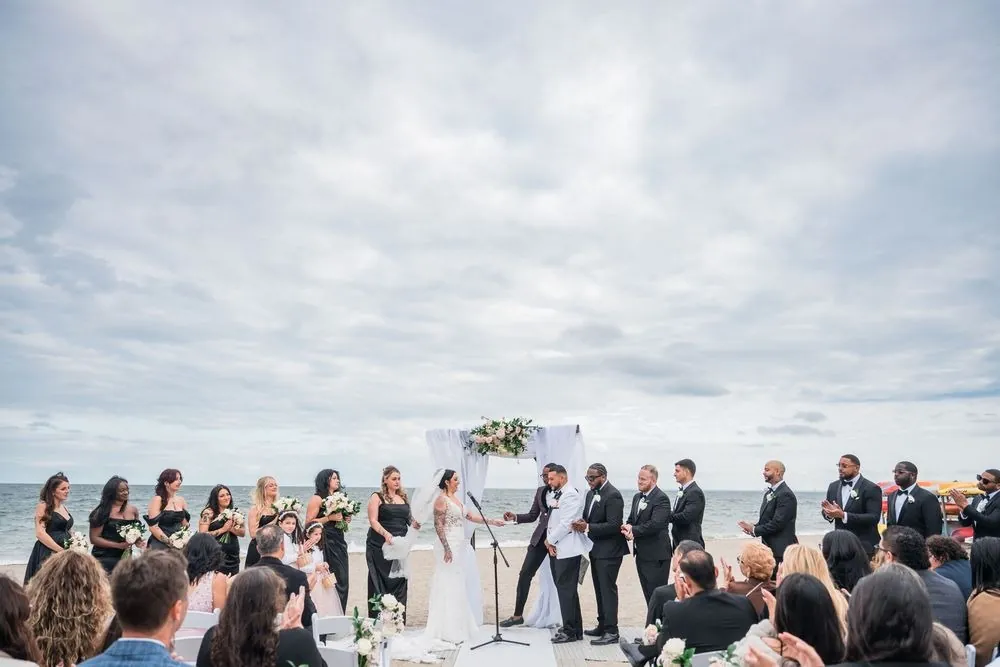 Wide view of the beach ceremony under heavy grey clouds, wedding party lined up beside the arch