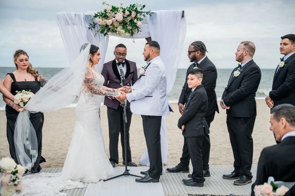 Olivia and Elvin hold hands under a draped arch with roses as the officiant reads, surf behind them