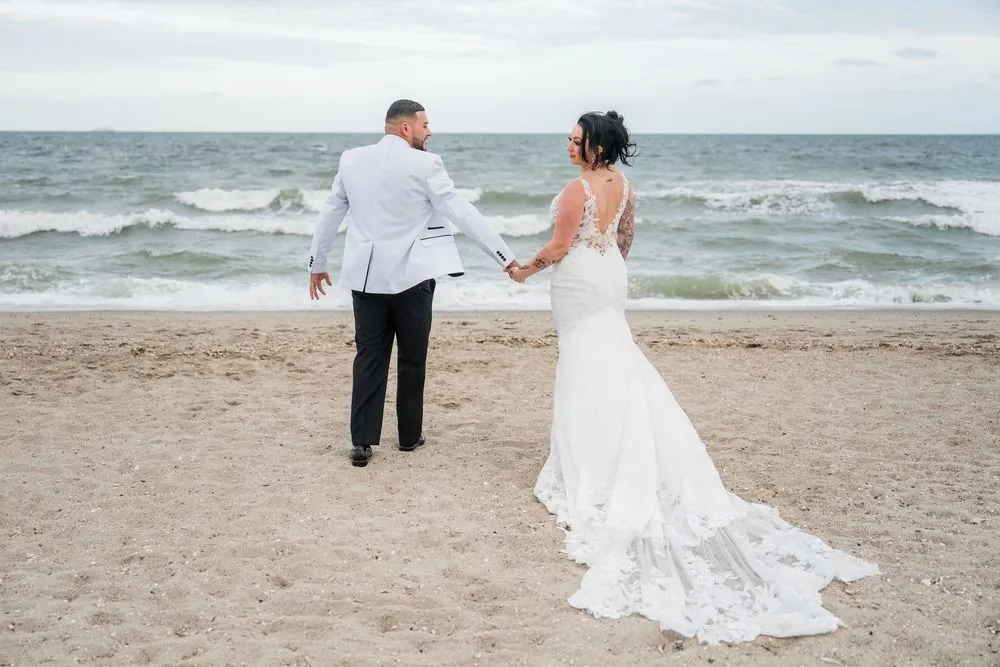 Olivia and Elvin walk hand in hand toward choppy surf, her lace train dragging across the sand