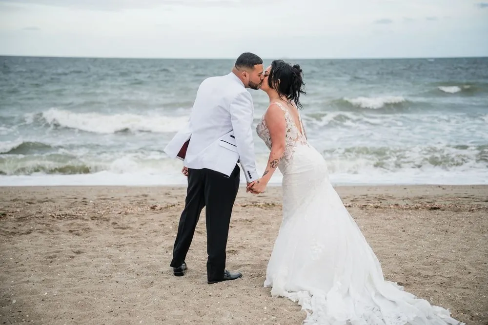 Olivia and Elvin kiss at the waterline under an overcast sky, waves breaking white behind them