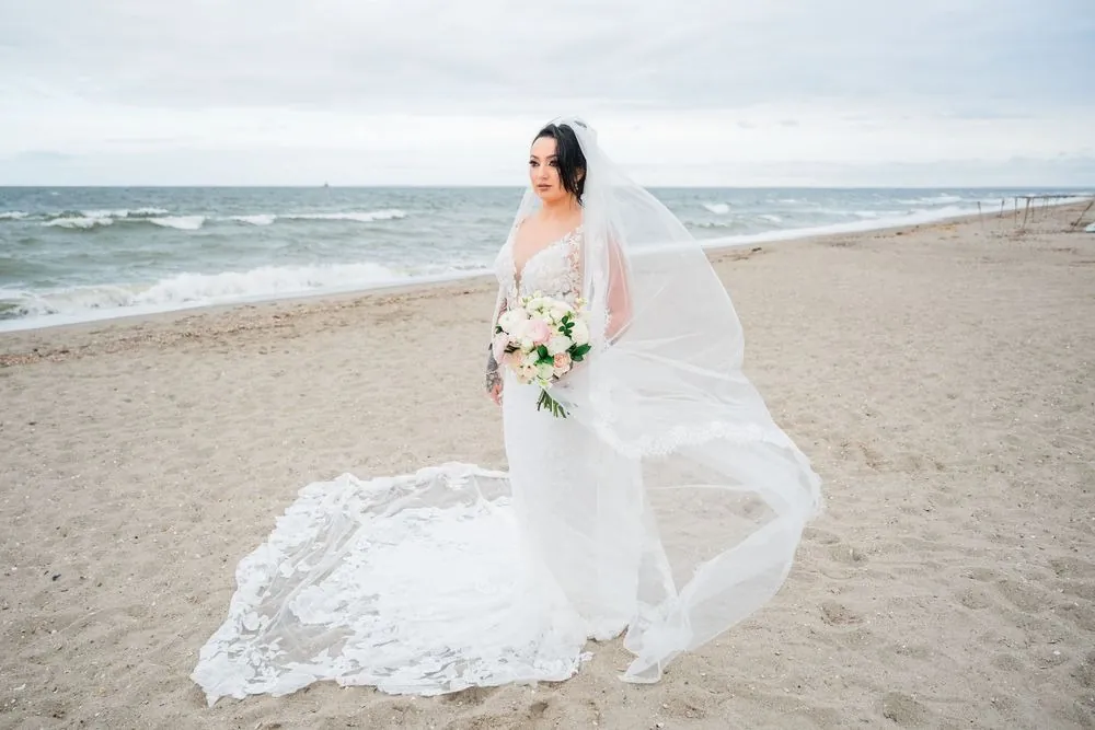 Olivia stands on the beach holding her bouquet while the wind lifts her long veil sideways