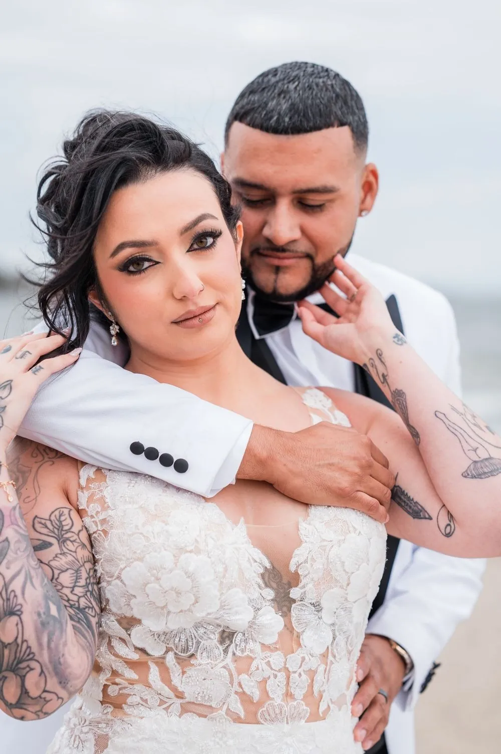 Elvin wraps his arms around Olivia from behind on the beach as she looks into the lens