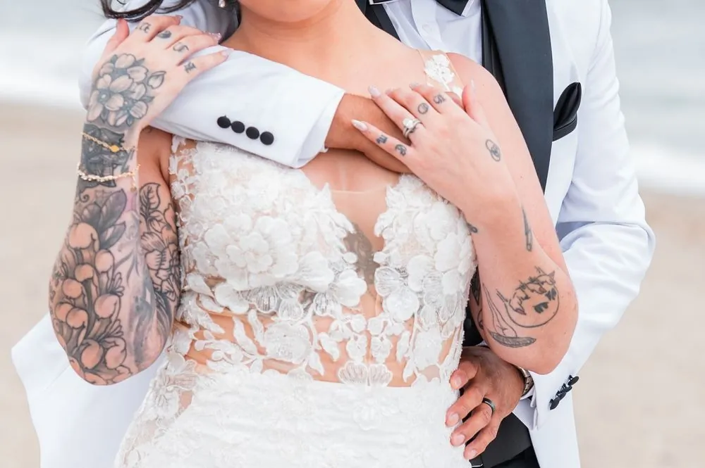 Close view of tattooed arms, rings, and a white jacket sleeve as the couple embrace on the beach