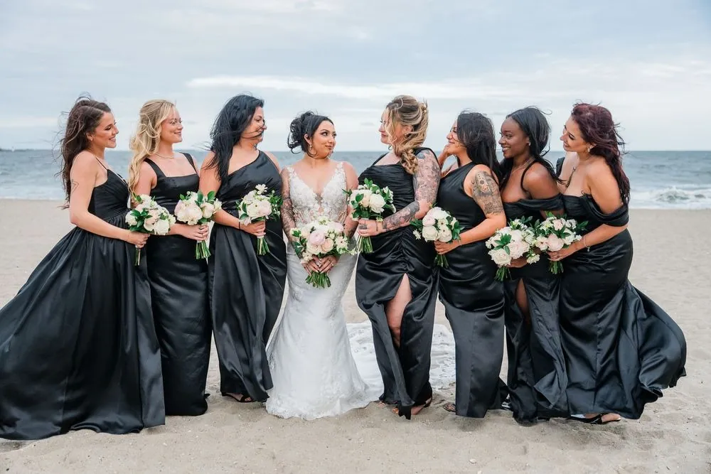Bridesmaids in black satin gowns laugh around the bride on the beach, bouquets in hand