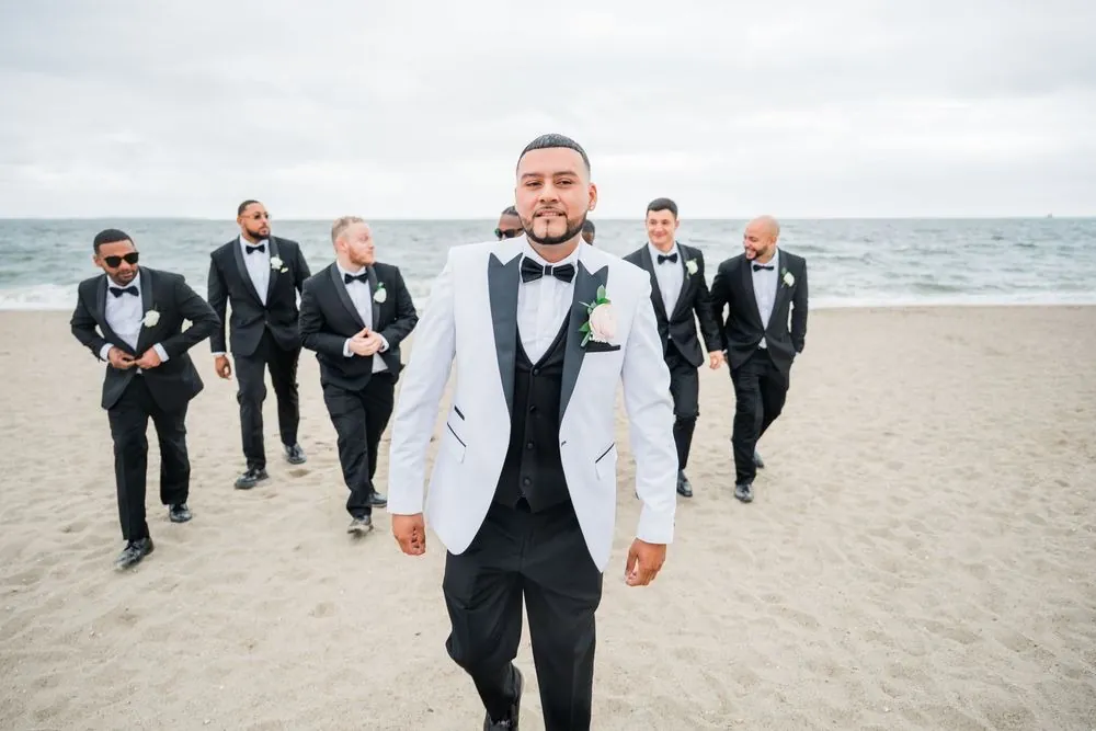 Groom in a white jacket strides across the sand ahead of his groomsmen in black tuxedos