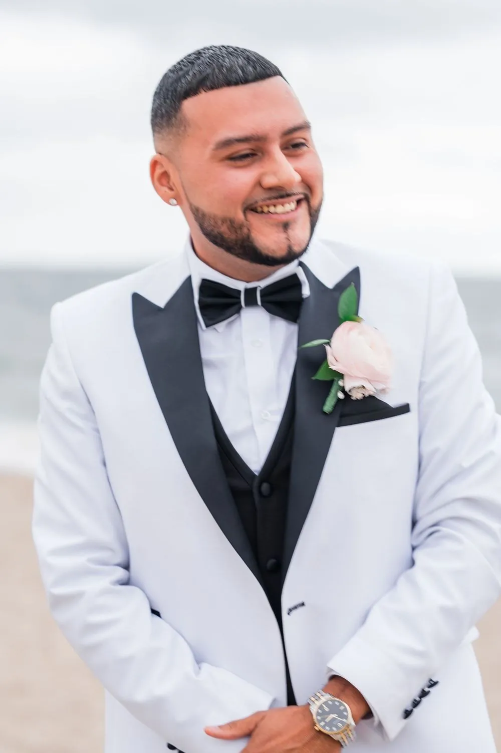 Groom smiles in a white dinner jacket with a blush rose boutonniere, the Sound behind him