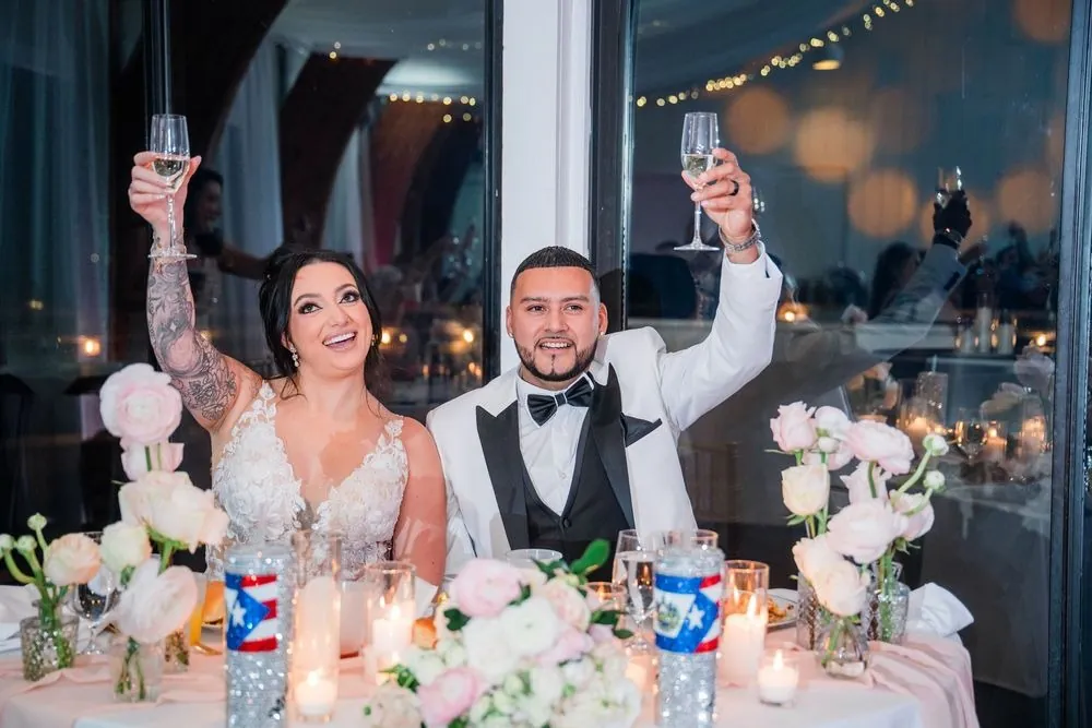 Olivia and Elvin raise champagne at a sweetheart table of blush roses and flag-wrapped glasses