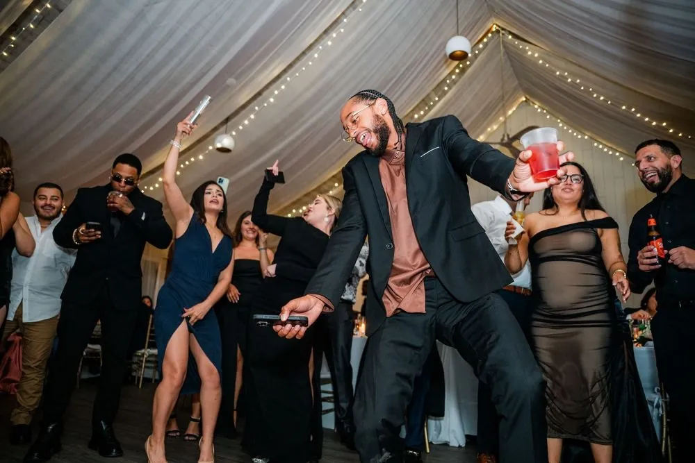 Guest in a black suit dances low with a red cup as friends cheer under string lights