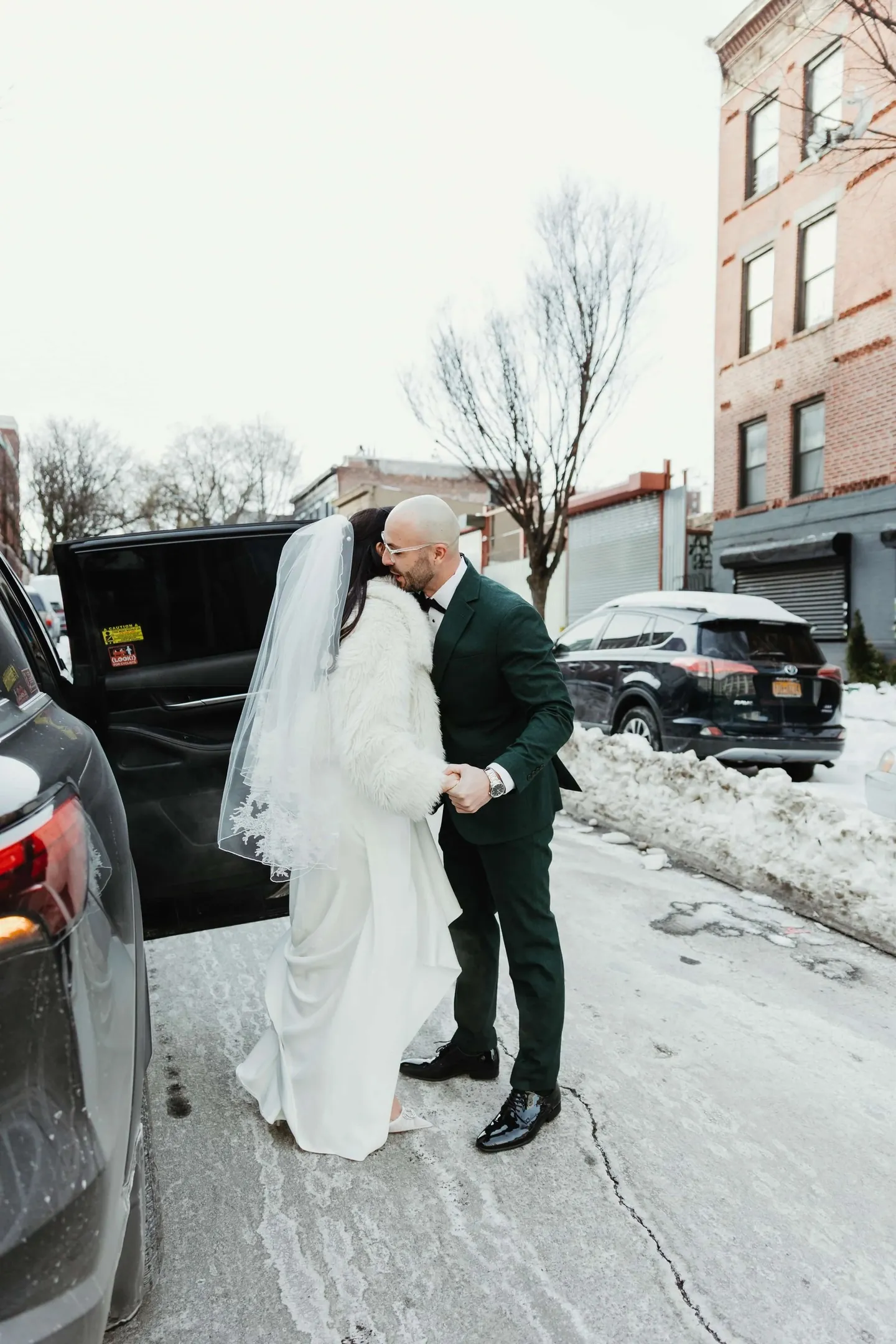 Rachel and Joe stand close on a snowy Brooklyn sidewalk between parked cars