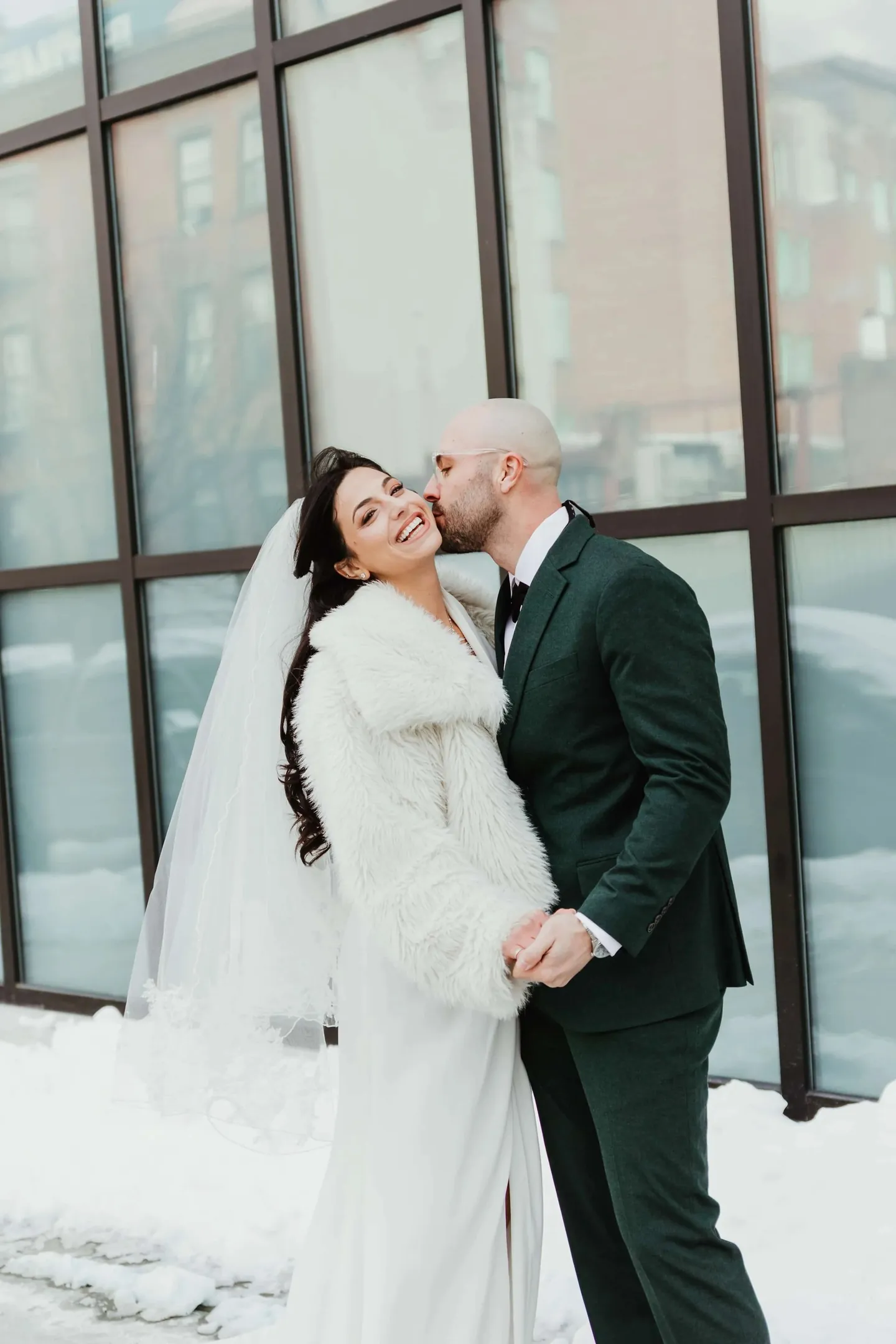 Rachel and Joe kiss in front of tall steel windows in soft daylight