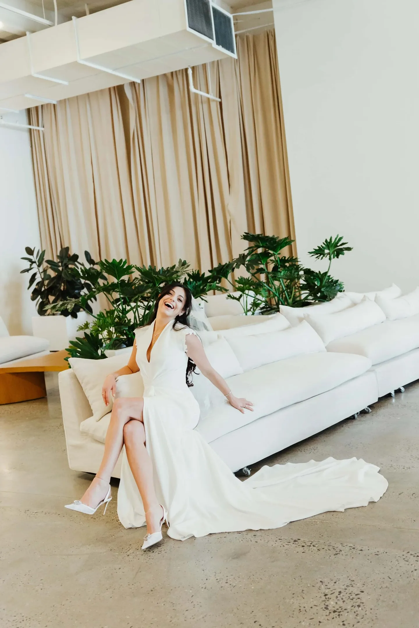 The bride lounges on a long white sofa in a bright room with potted greenery