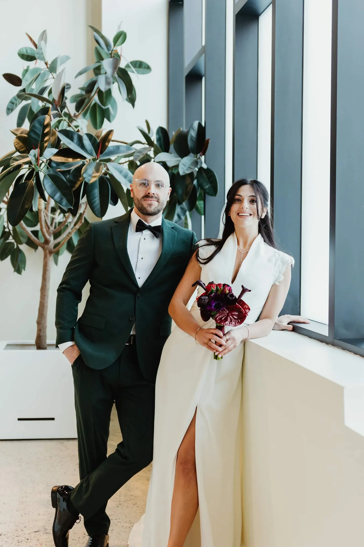 Rachel and Joe lean against a window ledge beside a tall potted tree