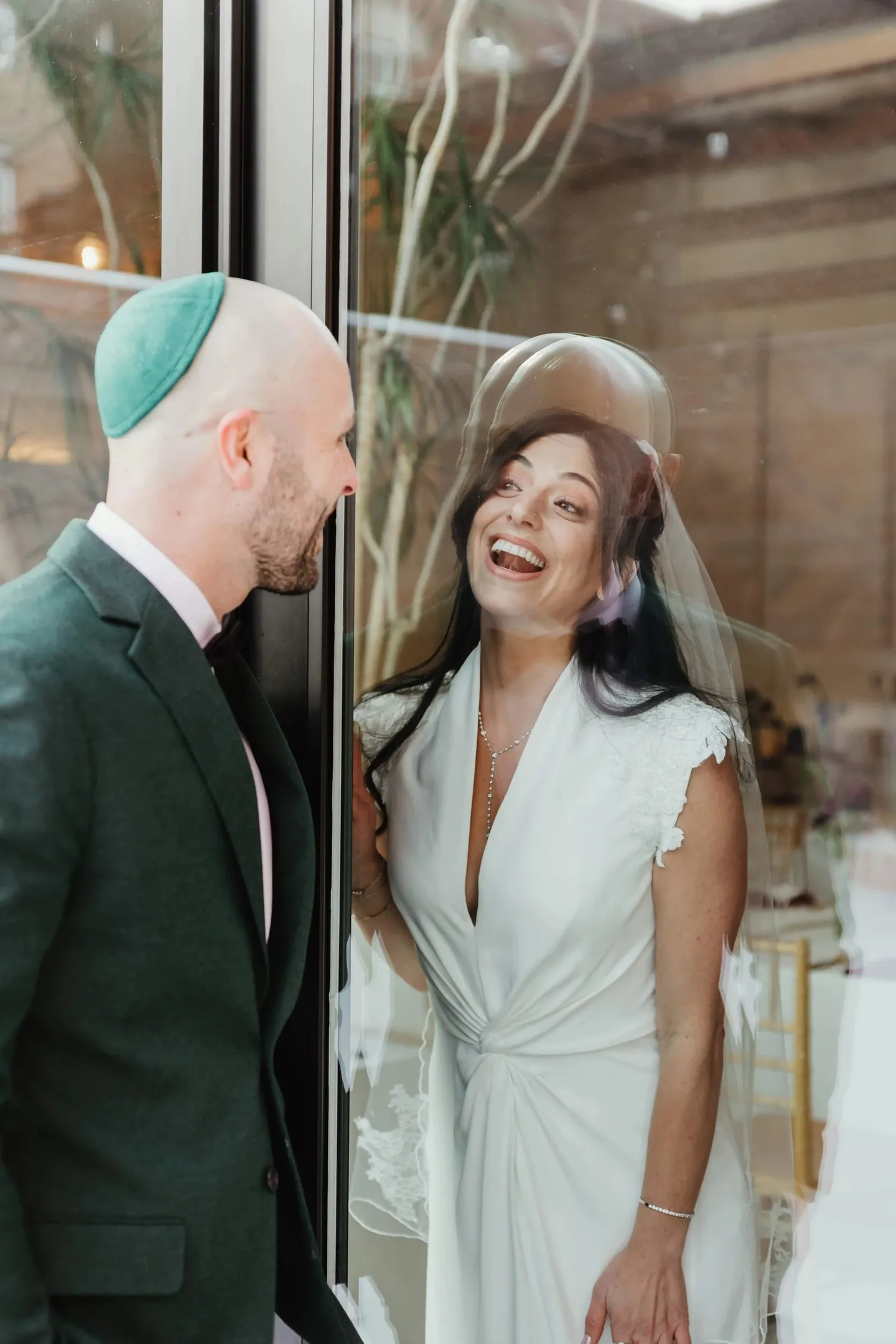 The bride laughs under her veil while the groom in a green kippah smiles at her