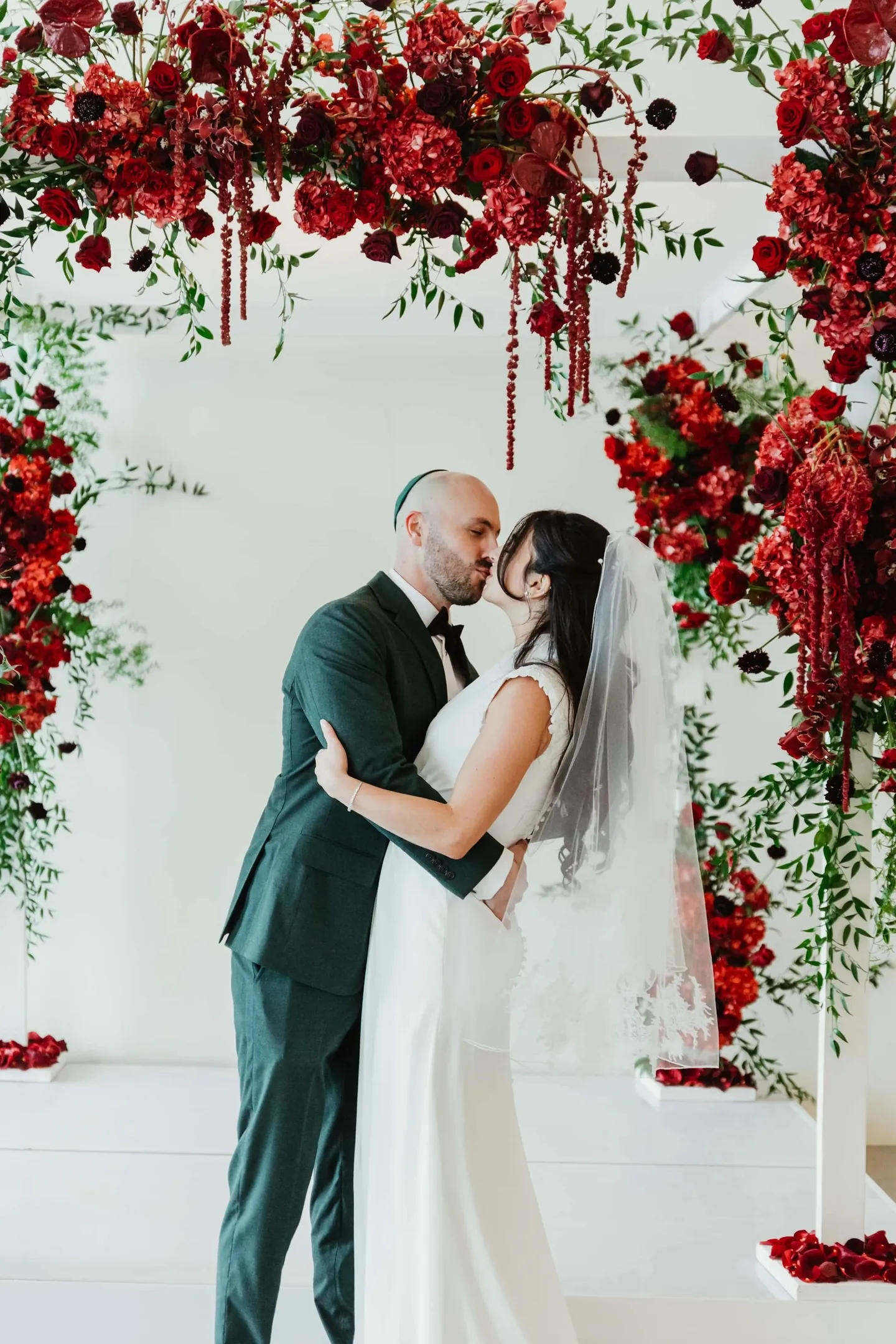 Rachel and Joe kiss beneath a chuppah covered in dark red roses and hanging amaranth