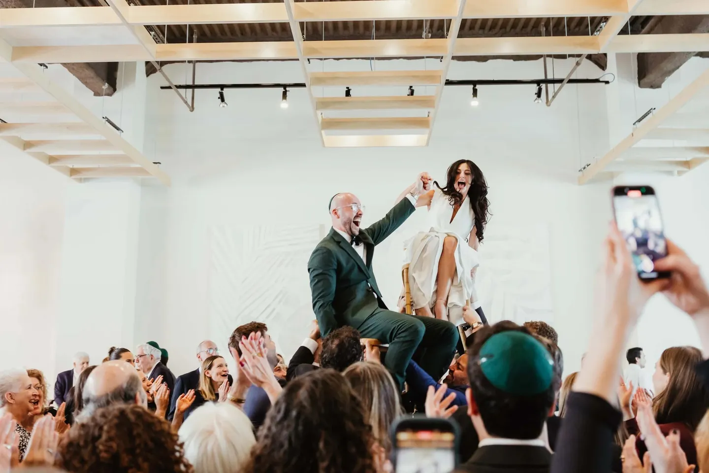 Rachel and Joe are lifted on chairs above a crowd of raised phones and kippot