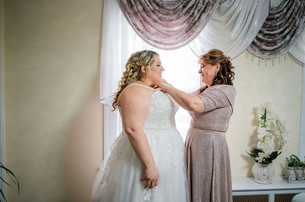 An older woman fastens the bride's necklace in front of a lace-curtained window