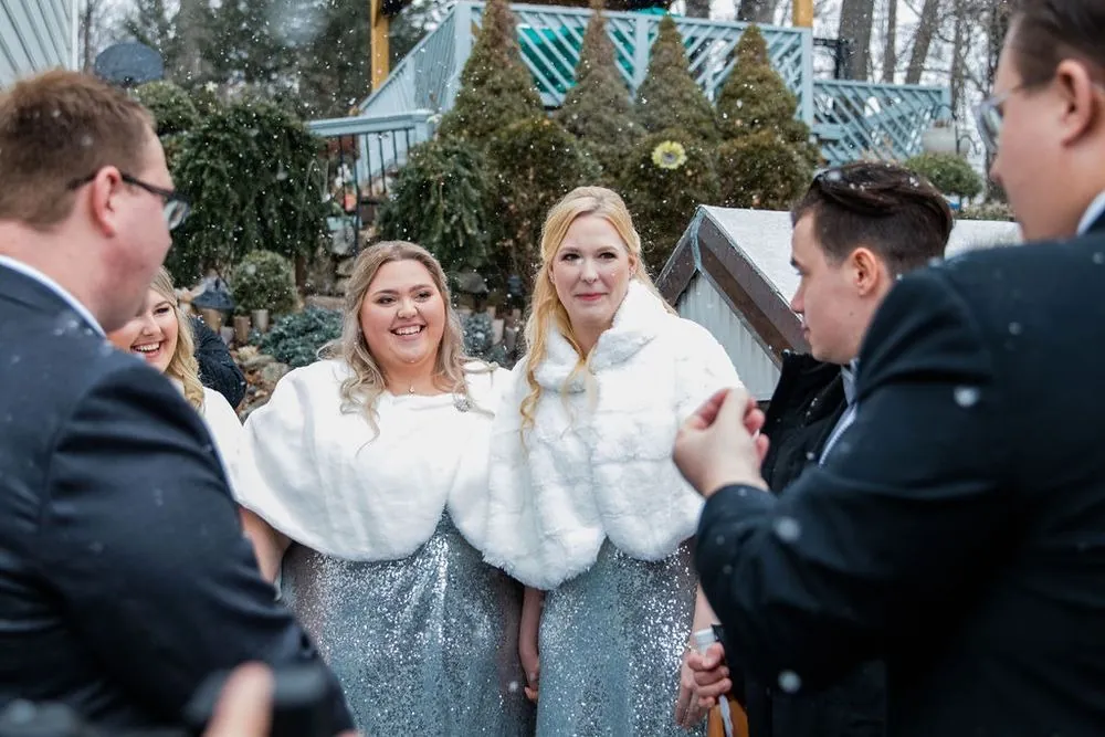 Bridesmaids in white faux-fur wraps and silver sequins laugh with groomsmen as snow falls