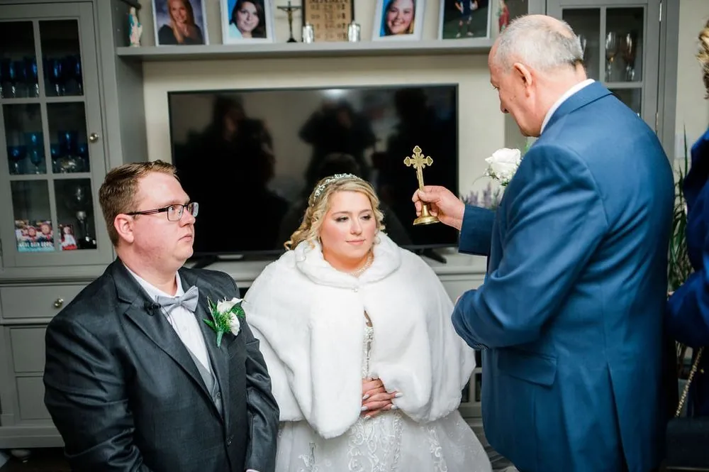 An older man in a blue suit holds a brass crucifix over the seated couple at home