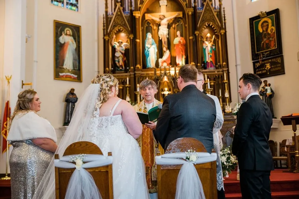 Sandra and Daniel stand before the priest at a carved altar as the wedding party looks on