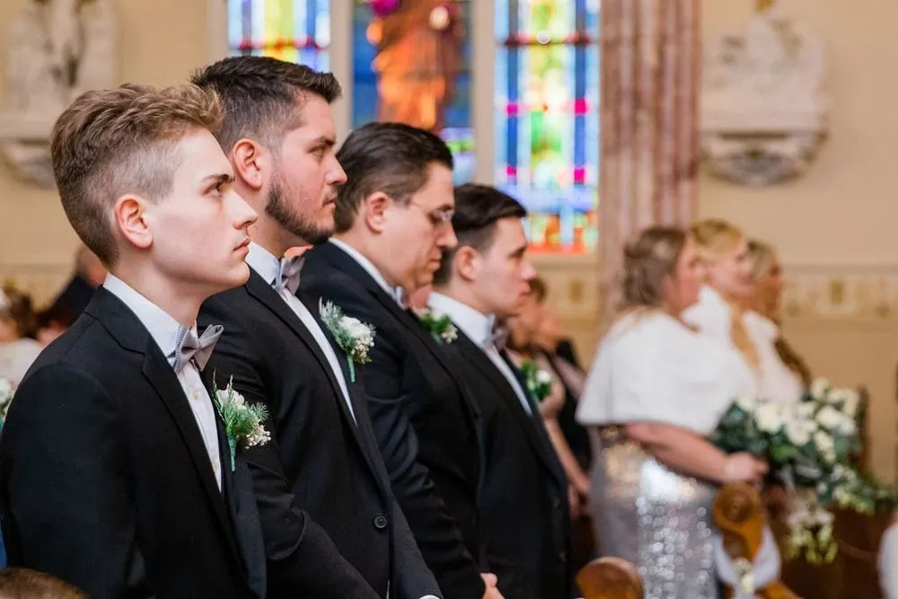 Groomsmen in black tuxedos watch the ceremony from the front row near stained glass