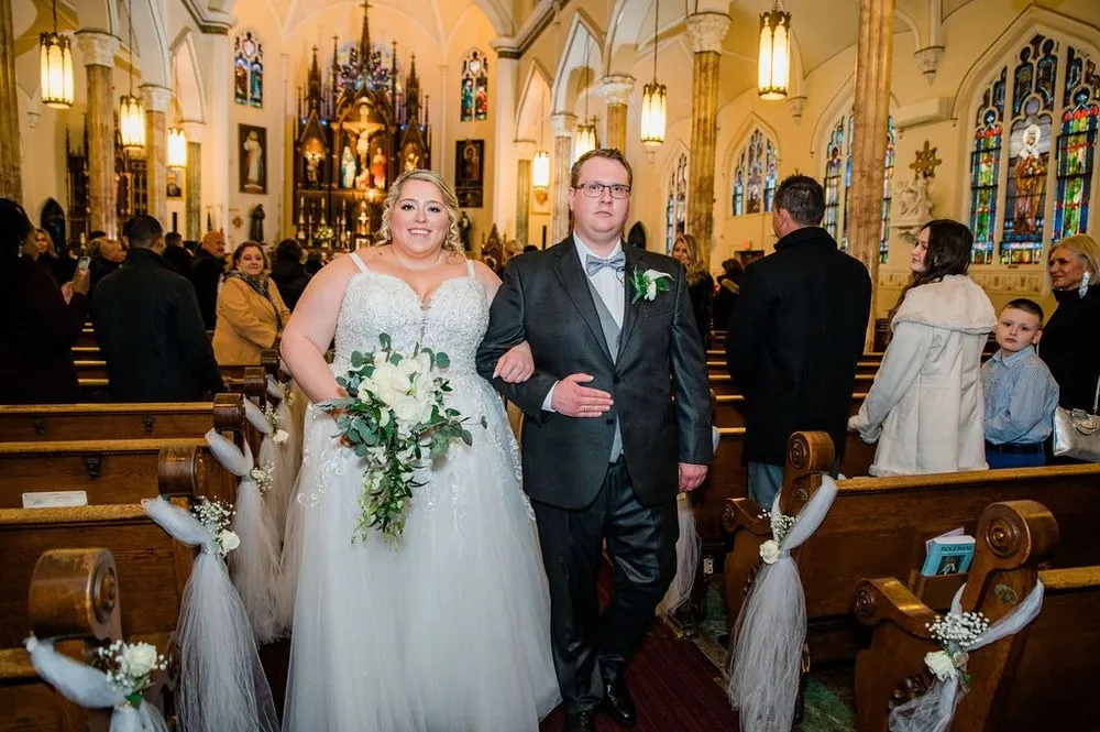 Sandra and Daniel walk arm in arm down the church aisle past pews tied with tulle