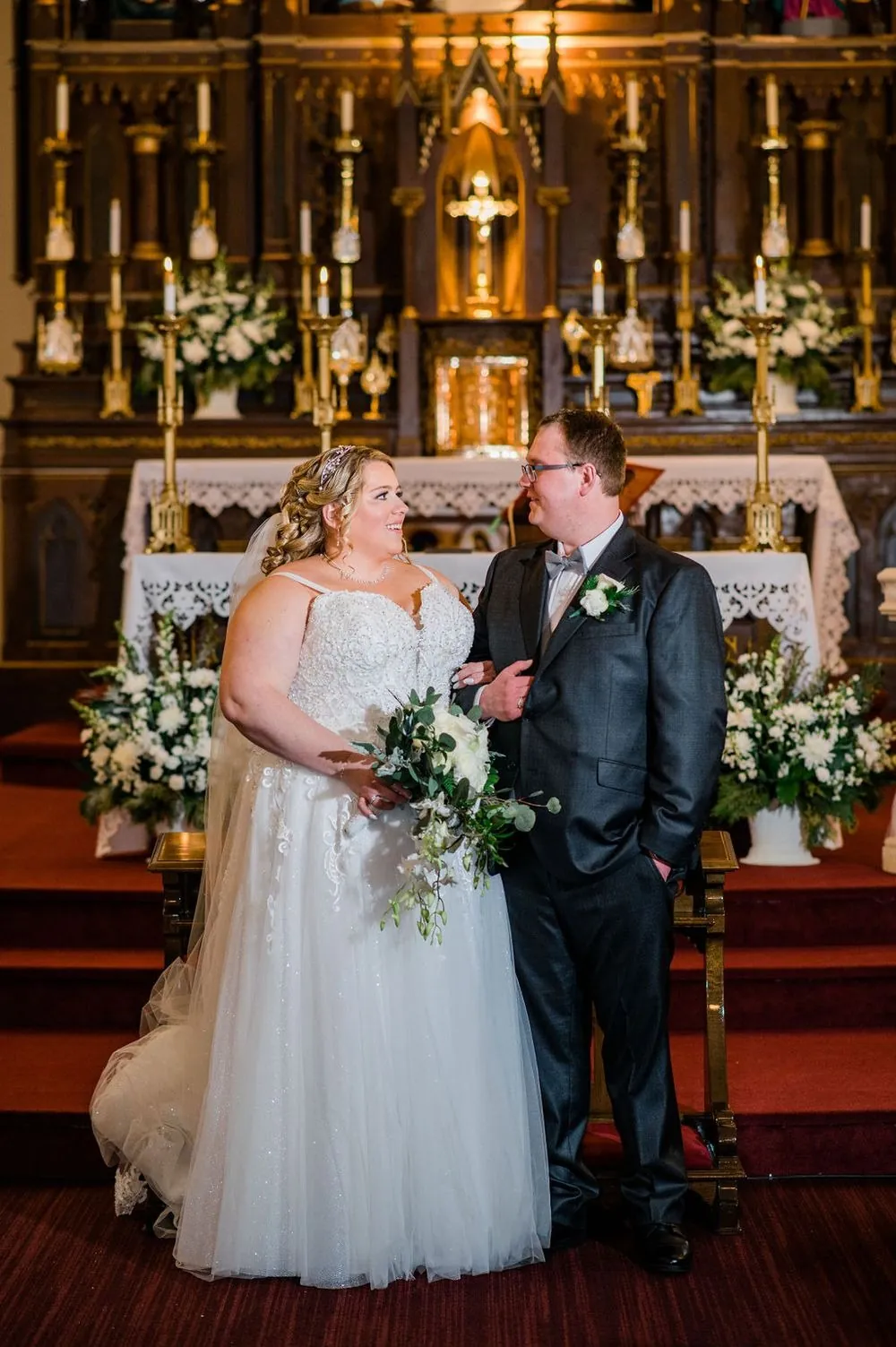 Sandra and Daniel pose in front of a golden altar with white flowers and candles