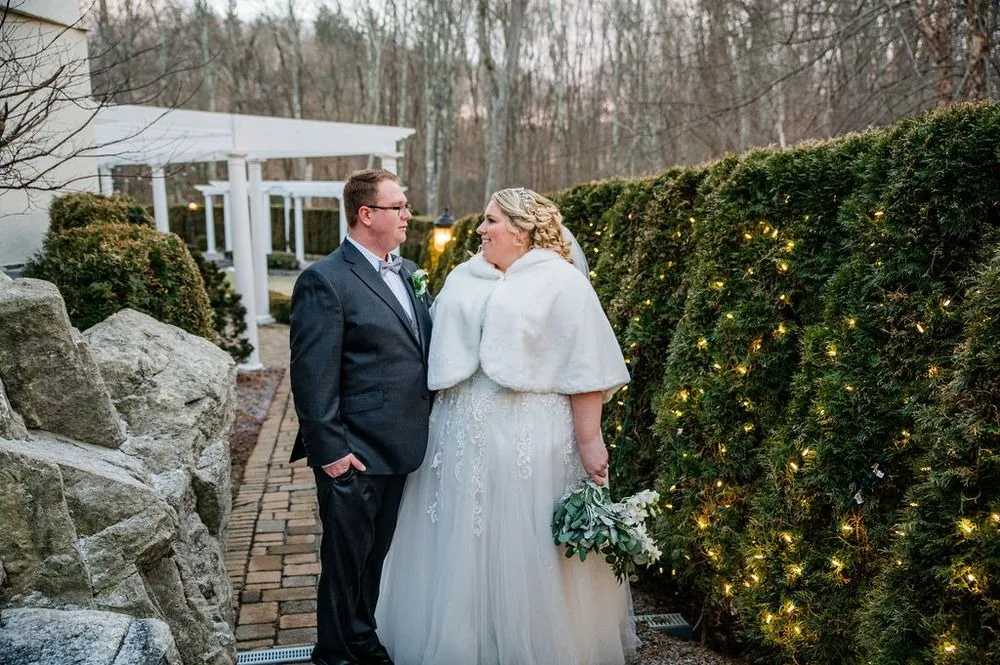 Sandra and Daniel stand on a brick path at Cascade beside hedges wrapped in glowing string lights