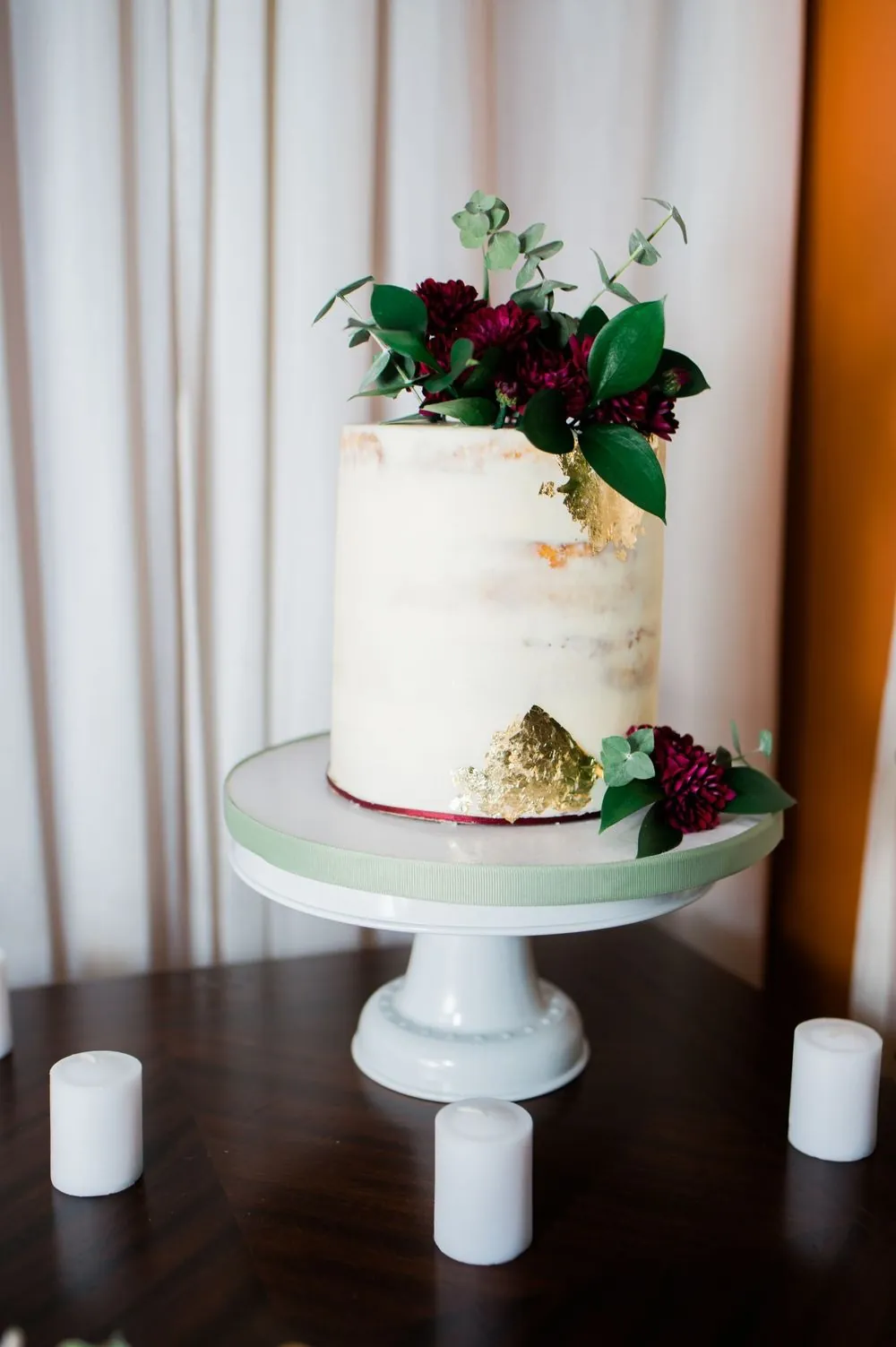 A naked cake topped with dark red flowers on a white stand
