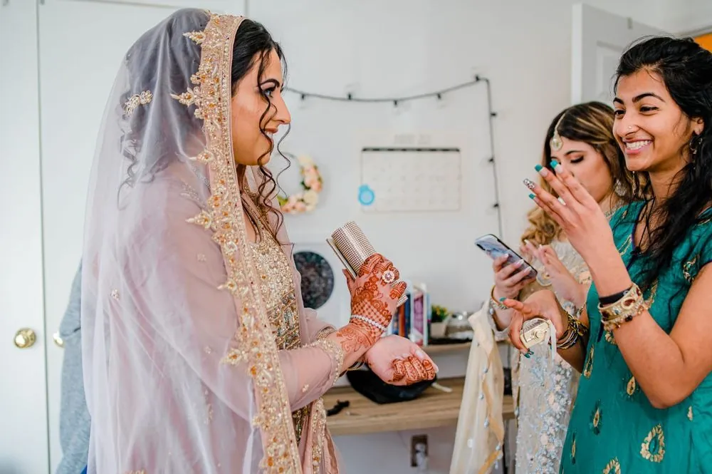 The bride shows her henna to laughing friends holding phones in her bedroom