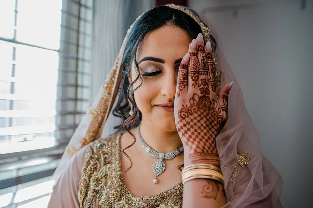 The bride touches her cheek with a hennaed hand beside a sunny window