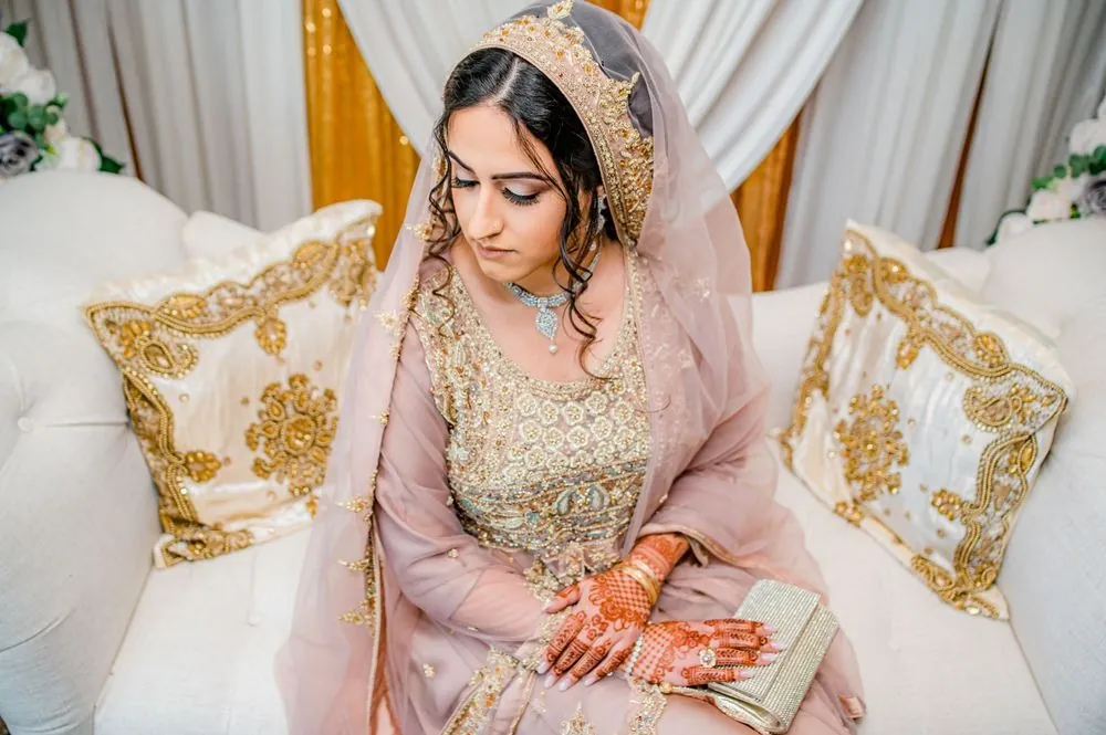 The bride sits on the white settee under the floral arch, hands folded in her lap
