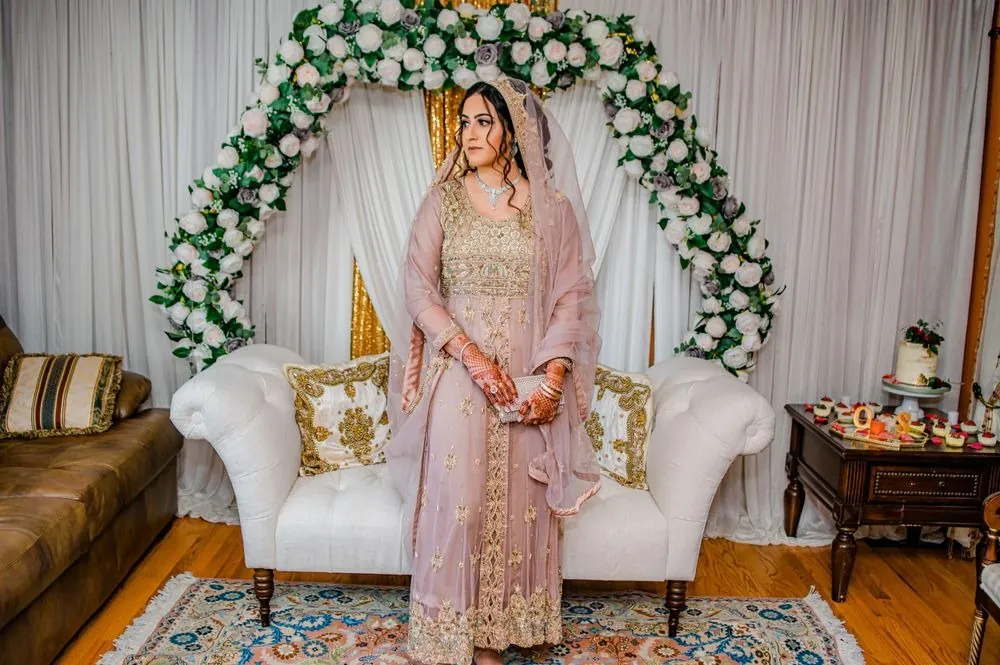 The bride stands before the rose arch in her blush gown and gold-trimmed dupatta