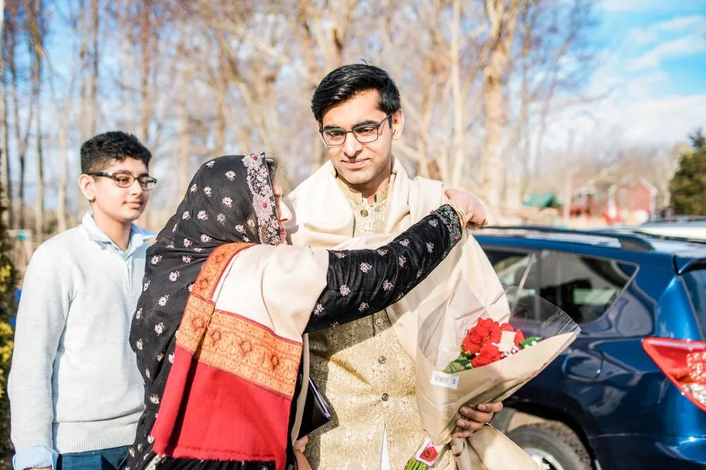 A woman in a red shawl hugs the groom as he arrives holding red roses