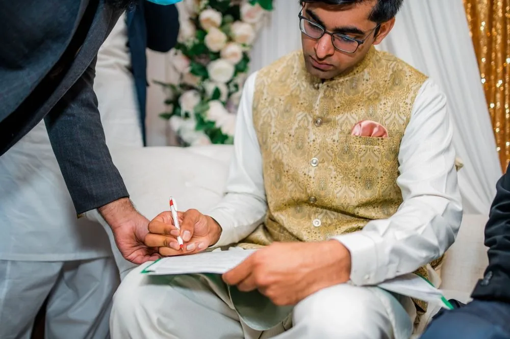 The groom signs the marriage papers seated in front of the floral arch