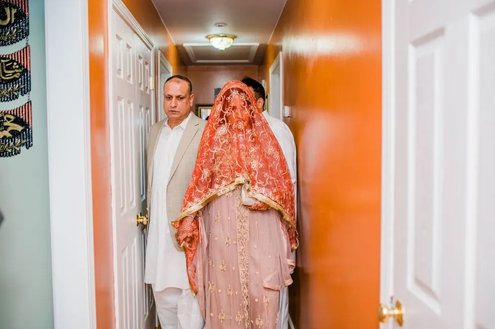 The bride walks down the hallway under an embroidered coral veil with family beside her
