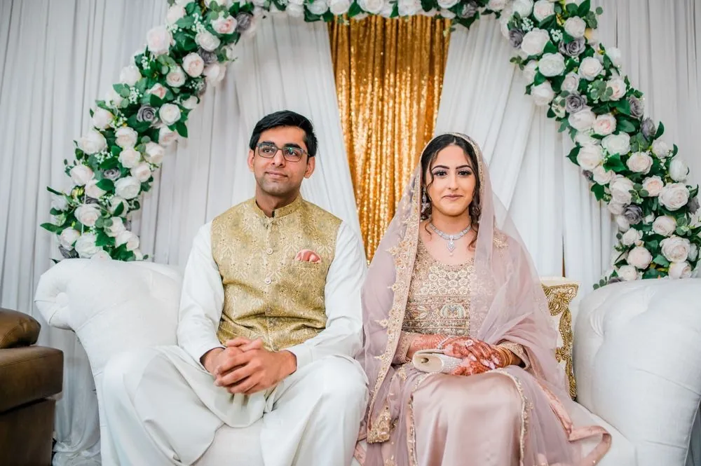 Sohaib and Qaniat sit side by side on the white settee beneath the arch