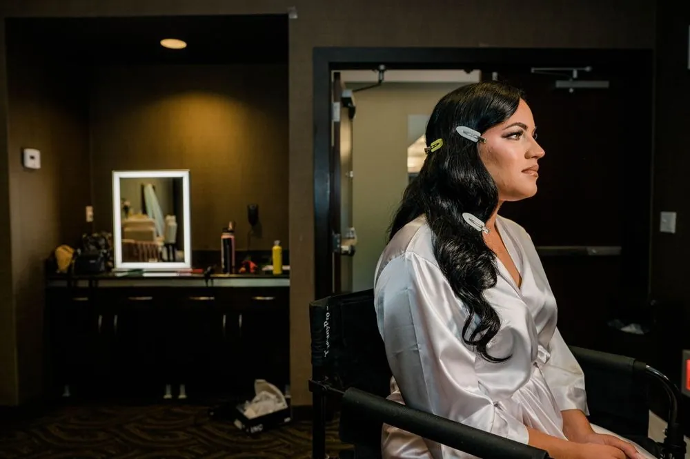 The bride sits in a satin robe beside a lit vanity mirror, a jeweled clip in her hair
