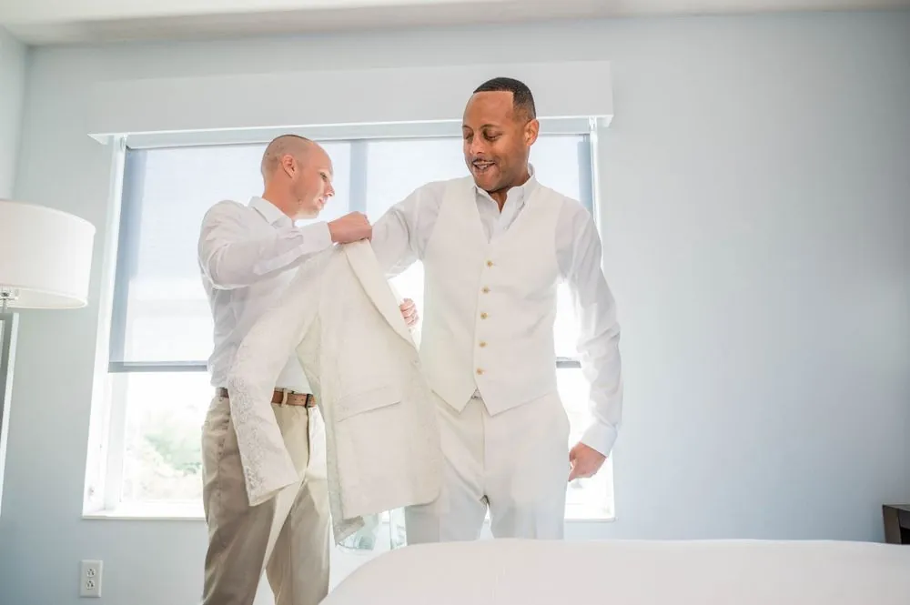 A groomsman helps the groom into his white jacket beside a bright hotel window